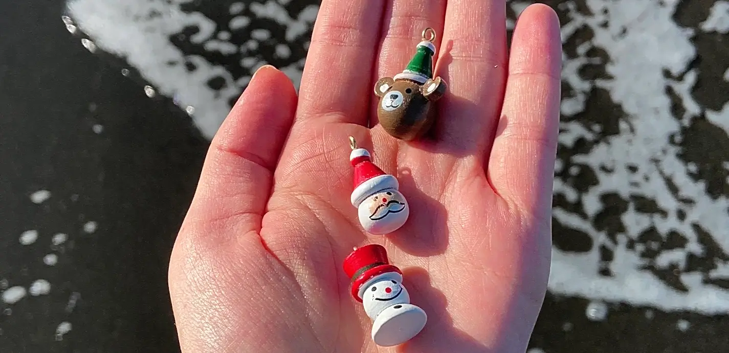 A person’s open hand holds three small painted Christmas ornaments against a background of dark wet sand and white ocean foam: a brown bear in a green-and-white hat, a Santa Claus face with red hat, and a snowman with a red top hat.