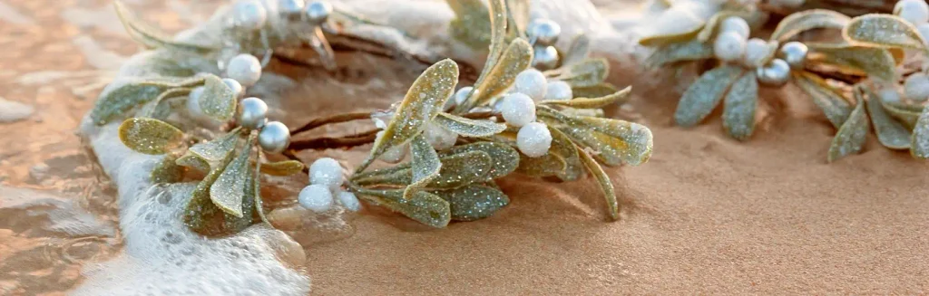 Close-up of a glittery mistletoe-style decoration with green leaves and white pearl-like berries resting on wet sand. Gentle ocean foam washes over the edge of the arrangement.