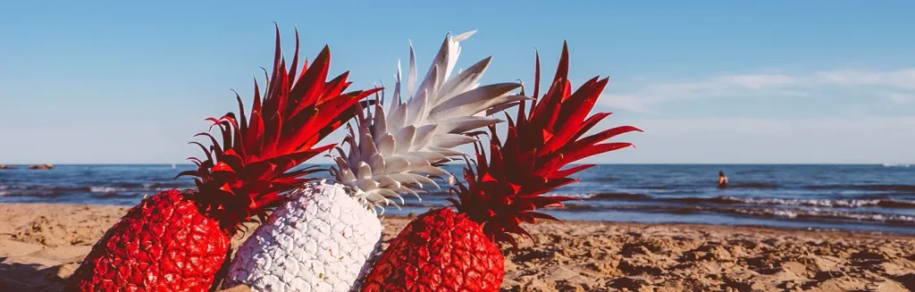 Three decorative pineapples sit on a sandy beach: two painted bright red and one painted white, all with spiky leafy tops. The blue ocean and clear sky form the background, with a few distant figures in the water.