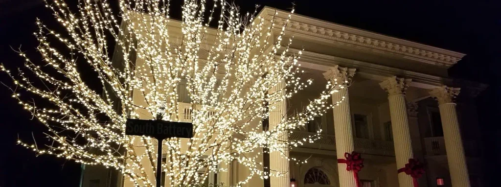 Nighttime view of a large white historic building with tall columns decorated with red bows. In the foreground, a tree is densely covered in warm white string lights. A street sign reading “South Battery” is visible.