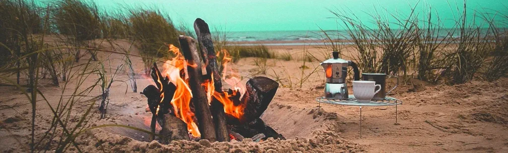 A small campfire burning among logs on a sandy beach with dune grass, next to a metal moka pot and two white cups on a small wire stand, with the ocean in the background