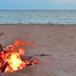 A small campfire made of driftwood burning on a sandy beach, with orange flames and glowing embers, and a calm ocean stretching to the horizon under a clear sky