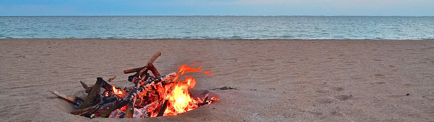 A small campfire made of driftwood burning on a sandy beach, with orange flames and glowing embers, and a calm ocean stretching to the horizon under a clear sky