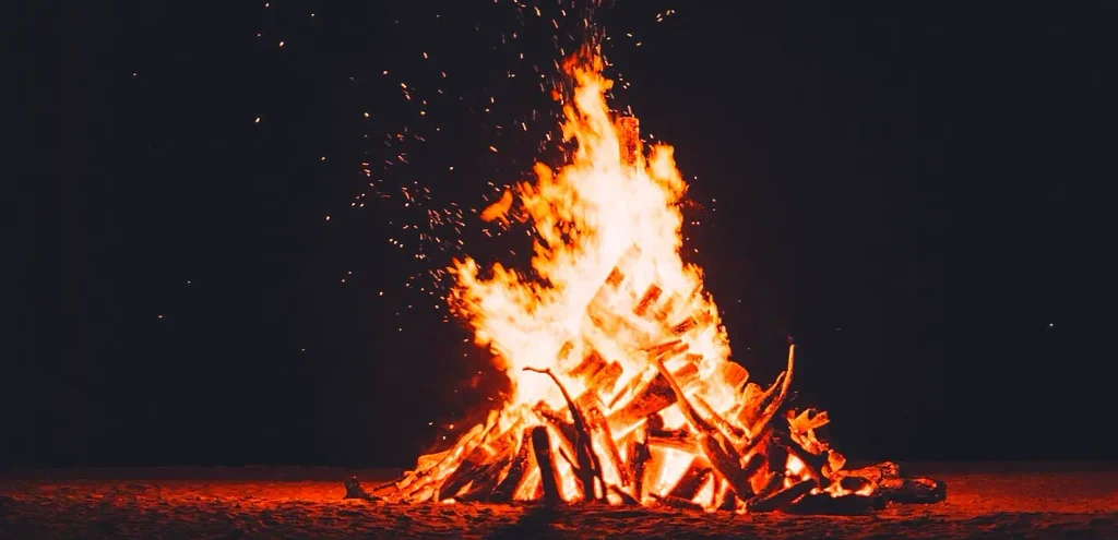 A large roaring bonfire with bright orange and yellow flames rising high against a dark night sky, sparks flying upward from a pile of burning logs on the ground