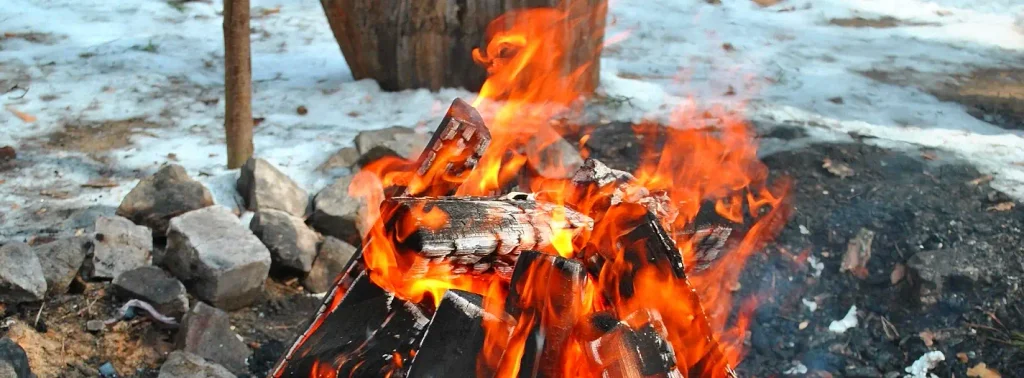 A bright campfire burning among charred logs, surrounded by gray rocks and patches of snow, with a wooden stump visible in the background
