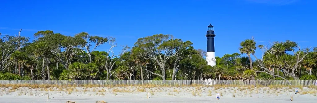 Tall black-and-white striped lighthouse rising above a line of green trees and palms, viewed from a sandy beach with a wooden fence in the foreground under a clear blue sky.