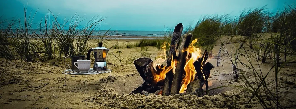 Small campfire of burning logs on the sand next to a portable stove holding a moka pot and white mug; dune grass and the ocean visible under a cloudy sky in the background.
