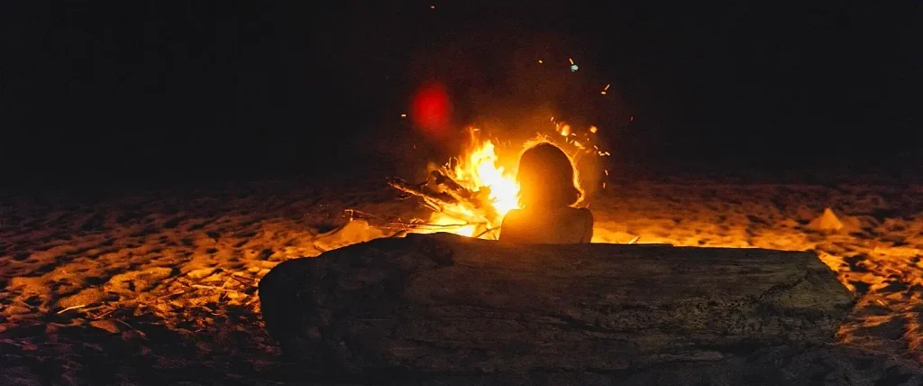 Person with shoulder-length hair sitting on a large driftwood log, back to the camera, facing a bright orange campfire with flying sparks against a dark night sky on the beach.