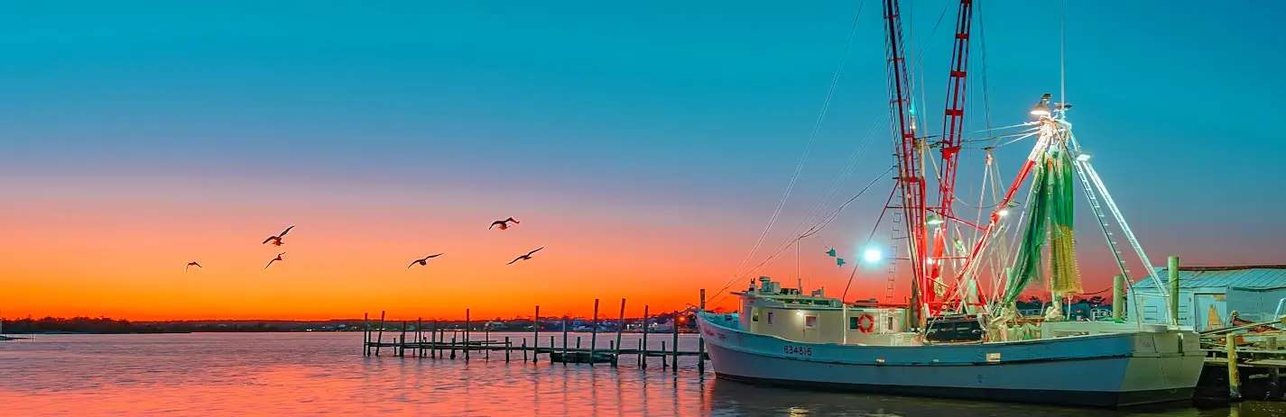 White fishing boat docked at a pier at dusk, with red masts and green nets, lights on, under a gradient sky of orange to deep blue, several birds flying left of the boat, calm reflective water.
