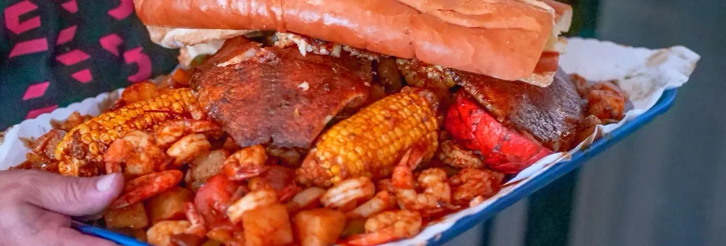 Close-up of a loaded seafood tray held by a hand: a sandwich piled high with fried seafood, corn on the cob, shrimp, potatoes, and a red crawfish, all coated in reddish seasoning.

