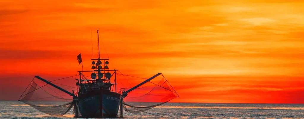 Fishing boat silhouetted against a brilliant solid orange sunset sky, outriggers and nets extended, facing the camera on calm water.

