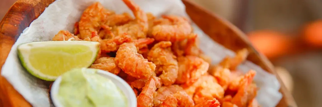 Basket of golden fried shrimp on white paper, with a lime wedge and a small cup of creamy green dipping sauce, in a wooden bowl.

