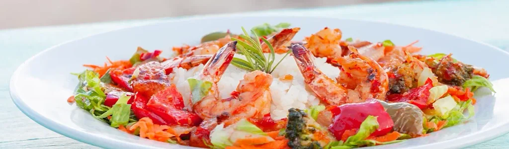 White plate of grilled shrimp arranged around a mound of white rice, mixed with red peppers, broccoli, carrots, and greens, garnished with a sprig of rosemary.

