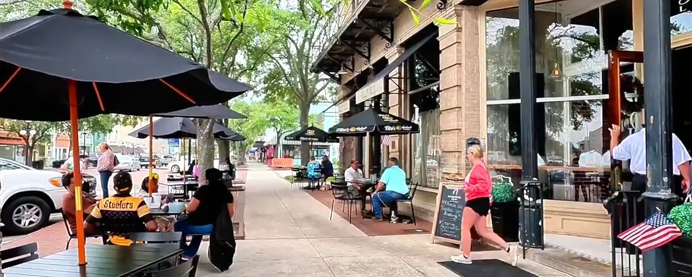 Sunny outdoor cafe seating area on a brick sidewalk with black patio umbrellas, people dining at tables (one wearing a Steelers jersey), a woman in a pink top walking past storefronts, and trees lining the street.