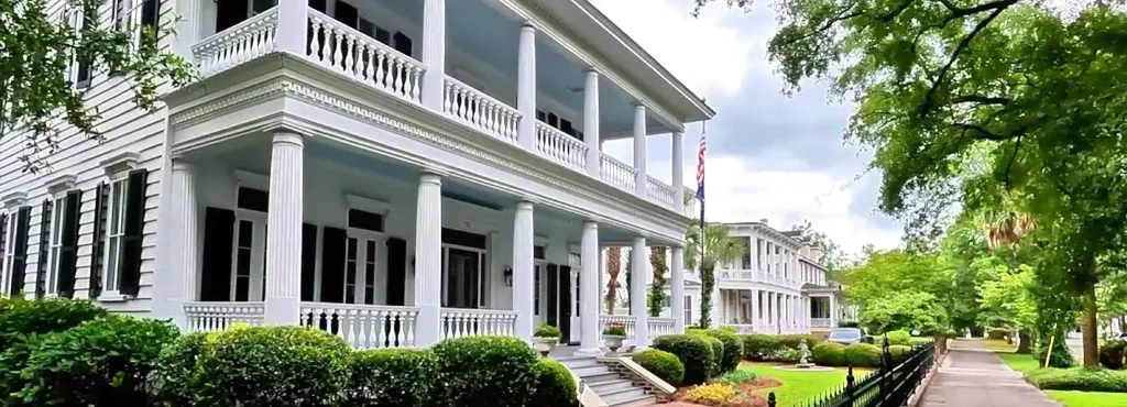 Large white two-story historic mansion with tall columns, wraparound balconies with white railings, black shutters, an American flag, manicured hedges, and a sidewalk under partly cloudy skies.

