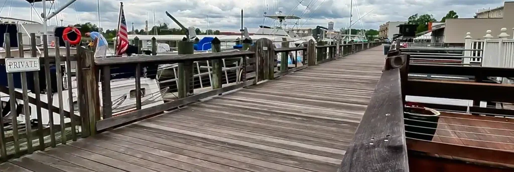 Wooden marina dock with railings, docked boats, an American flag, a “PRIVATE” sign, a life ring, and a person in a blue shirt near the boats under cloudy skies.

