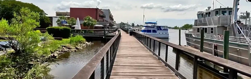 Long wooden boardwalk over calm water with metal railings, boats docked on the right, buildings and greenery on the left, under a partly cloudy sky.

