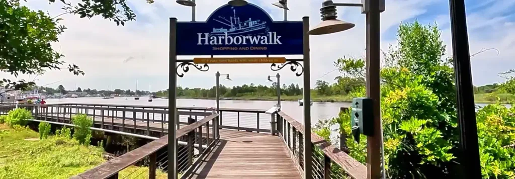 Entrance to a wooden boardwalk over water marked by a blue “Harborwalk Shopping and Dining” sign with a boat illustration, trees and greenery on both sides, and boats in the distance.

