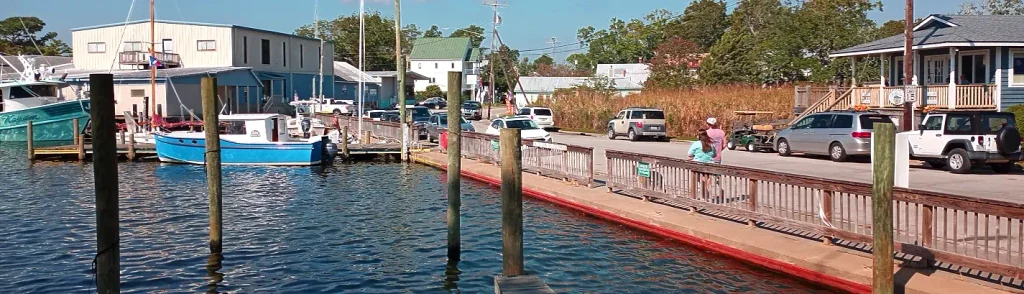 Sunny waterfront scene looking across a dock with wooden pilings toward parked cars, people walking, and buildings, with a blue-and-white boat named “Kayden” and other vessels on the left.

