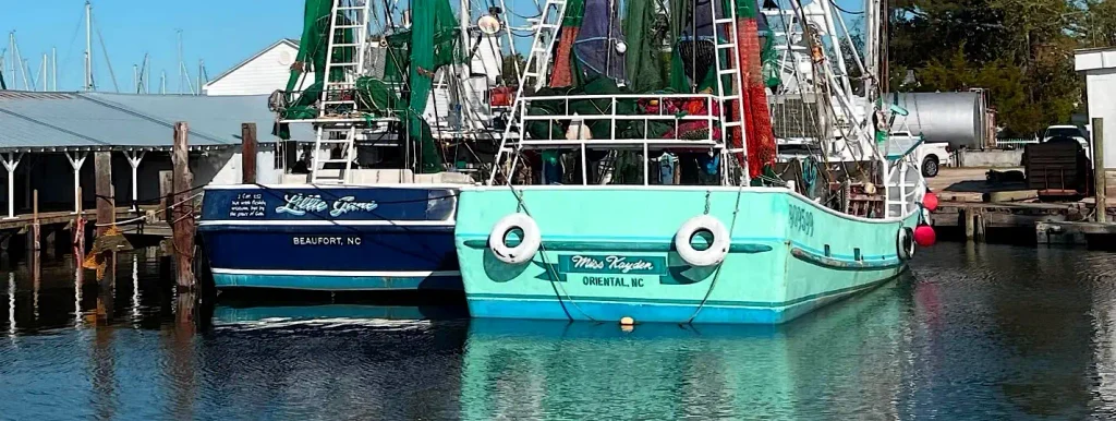 Sterns of two commercial fishing boats docked side by side: a dark blue boat named “Little Game” from Beaufort, NC, and a turquoise boat named “Miss Hayden” from Oriental, NC, with fishing nets visible.

