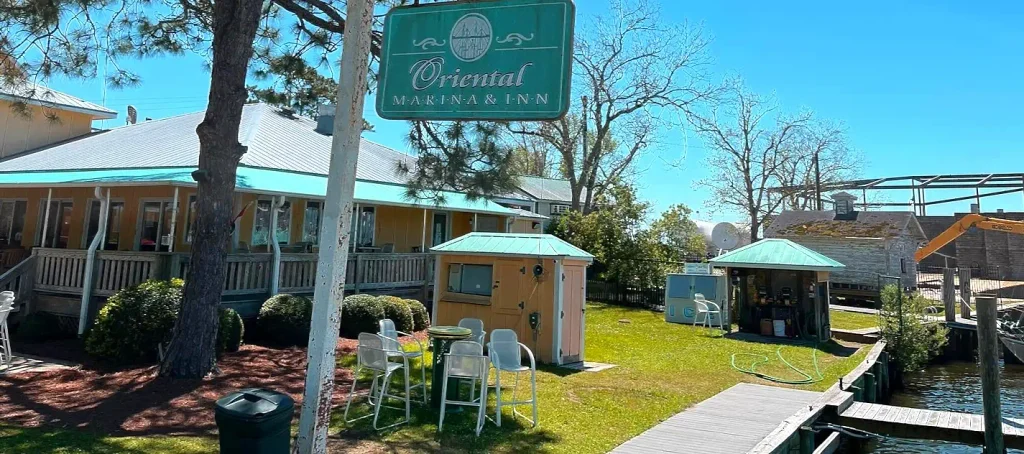 Teal “Oriental Marina & Inn” sign hanging from a white post in front of a yellow building with turquoise roof trim, outdoor seating, a small yellow shed, green lawn, and a dock on the right under a clear blue sky.

