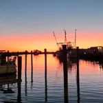 Silhouetted boats and docks in a marina at sunset, with the sky glowing orange and pink above calm reflective water.
