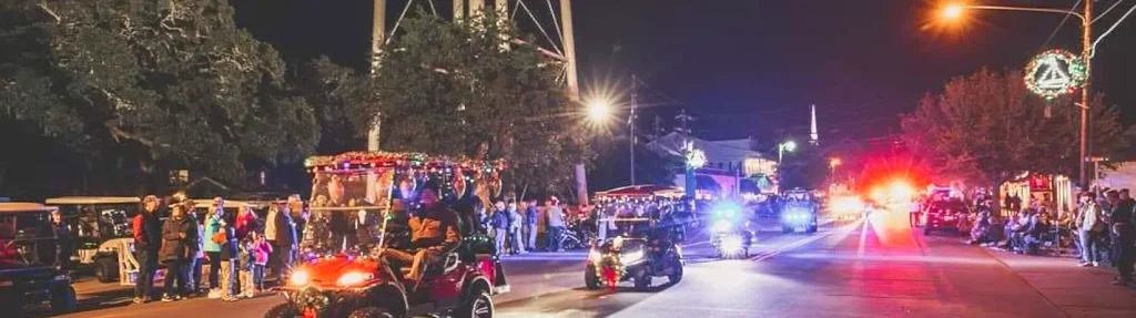 Nighttime Christmas parade featuring decorated golf carts and ATVs with colorful lights. Crowds of people line the street under streetlights and a water tower, with emergency vehicle lights flashing in the distance.