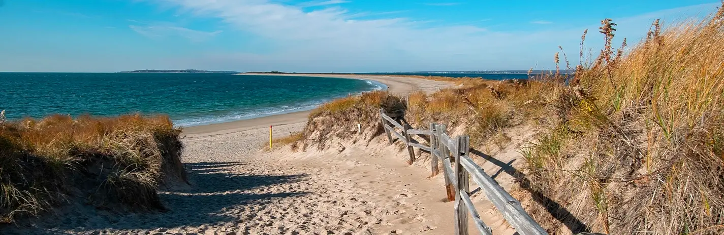 Sandy path leading down to a wide sandy beach and calm blue-green ocean, with wooden rail fence on the right, dry dune grasses on both sides, and distant land visible across the water under a clear blue sky.