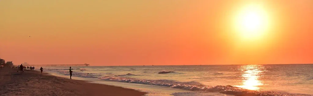 Vibrant orange and yellow sunset over the ocean with the sun low on the horizon reflecting on the water; people walking along the shoreline near a long wooden pier on the left, gentle waves rolling in.

