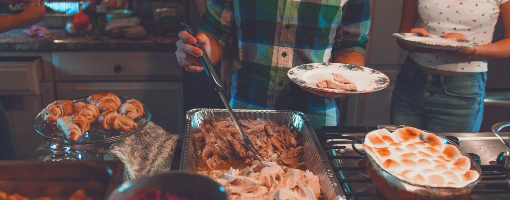 Kitchen scene of people serving food: a person in a green plaid shirt using tongs on a large tray of shredded turkey or roast, others holding plates, with a plate of croissants, a foil tray of meat, and a baked dish topped with toasted marshmallows in the foreground.

