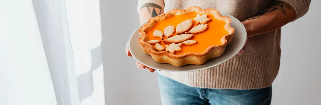 Person in a beige sweater and jeans holding a white plate with a whole pumpkin pie featuring a fluted crust and decorative leaf-shaped pastry cutouts on the bright orange filling.

