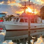 Calm marina at sunset with the white fishing boat “Miss April” docked in the center, its reflection clear in the still water. Other boats and waterfront houses line the background under a colorful sky of orange, yellow, and blue clouds.
