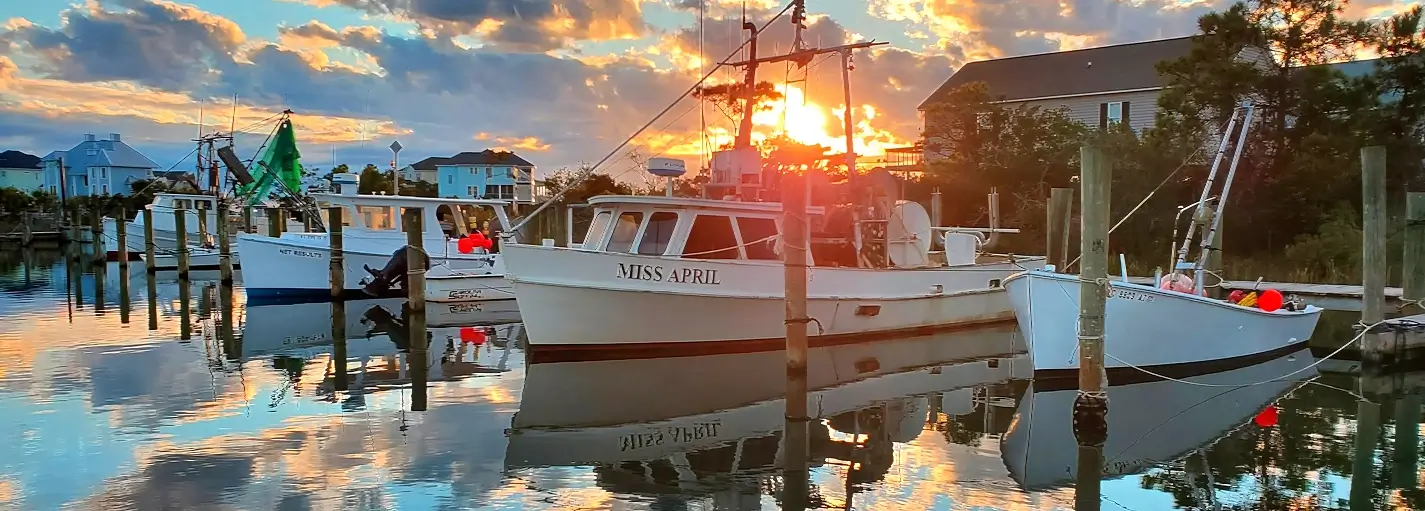 Calm marina at sunset with the white fishing boat “Miss April” docked in the center, its reflection clear in the still water. Other boats and waterfront houses line the background under a colorful sky of orange, yellow, and blue clouds.