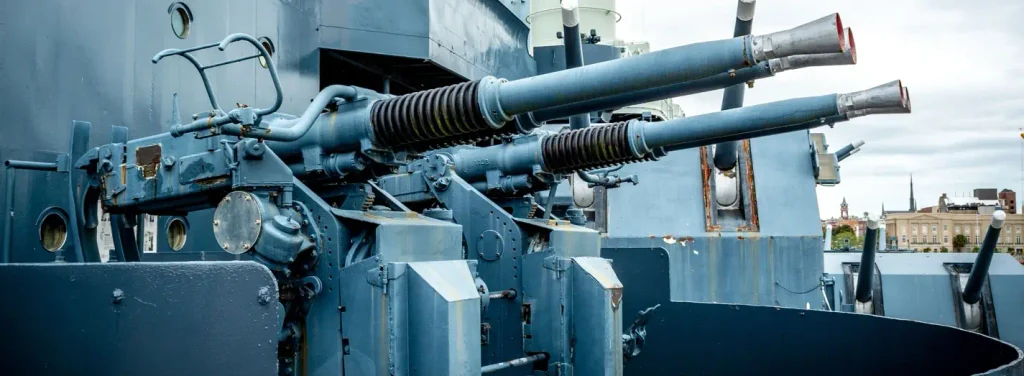 Close-up of dual-mounted gray naval anti-aircraft guns on a ship’s deck, showing the barrels, mechanical details, and a city skyline with a large building in the background under an overcast sky.

