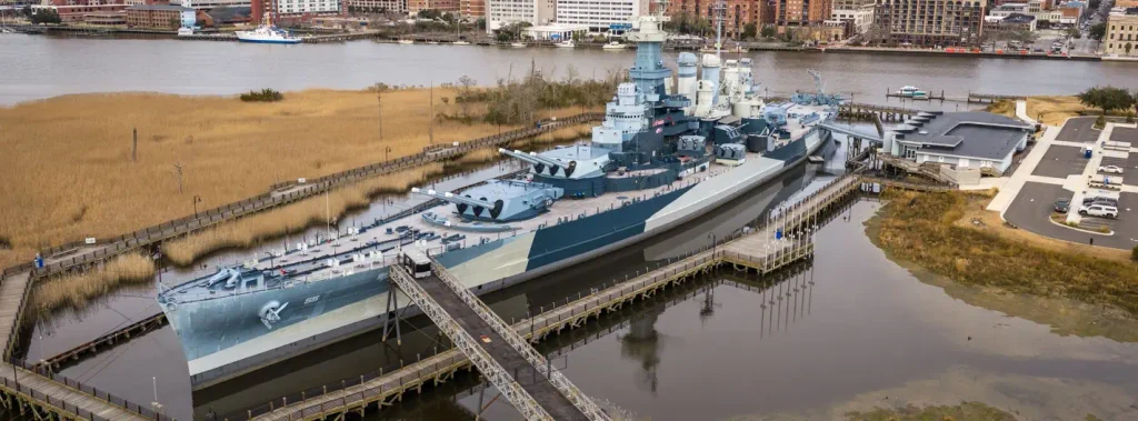 High-angle aerial photograph of a large gray battleship (BB-55) moored in a marshy waterway, connected by wooden walkways, with city buildings and a parking area visible nearby.

