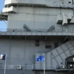 Side view of the gray superstructure of aircraft carrier “10” with two large American flags flying in the foreground and smaller flags between them under a clear blue sky.