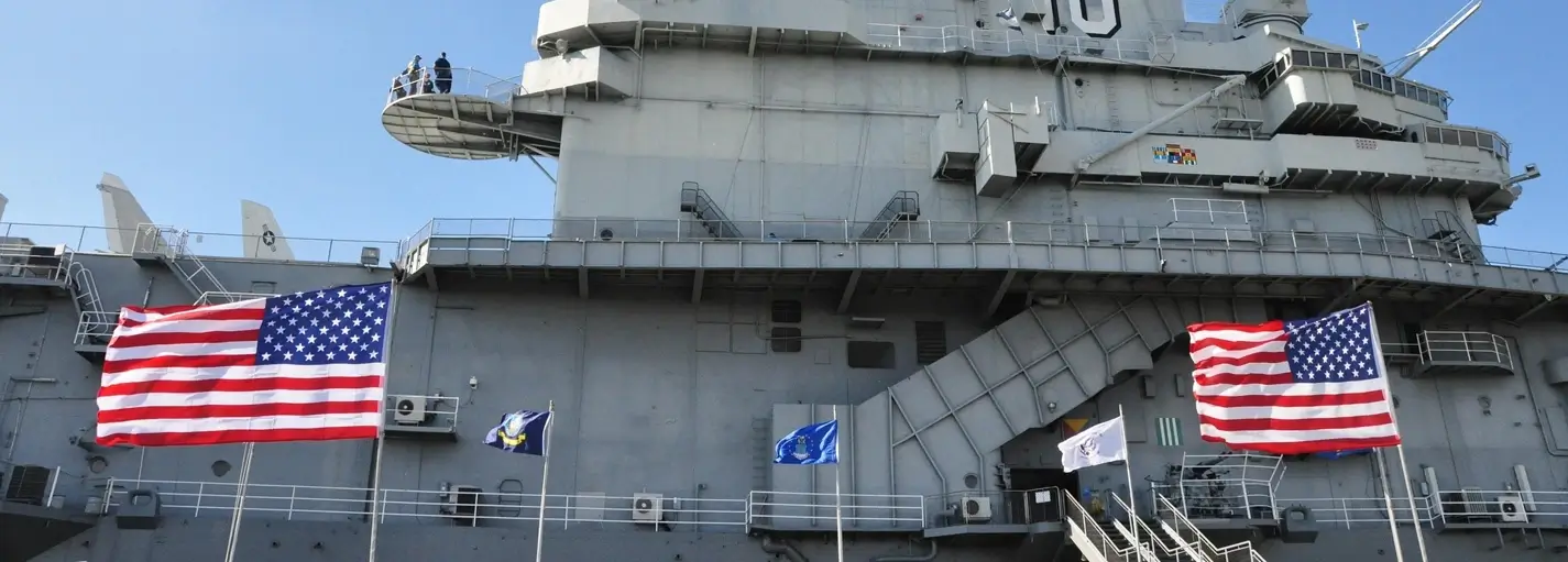 Side view of the gray superstructure of aircraft carrier “10” with two large American flags flying in the foreground and smaller flags between them under a clear blue sky.