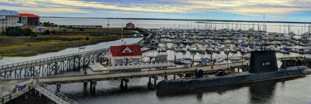 Wide view of a marina filled with sailboats and powerboats at sunset or sunrise, with a black submarine (hull number 343) docked on the right, a red-roofed building, boardwalks, and marshland in the background.

