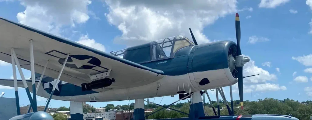 Blue-and-white World War II-era U.S. Navy aircraft (torpedo bomber/dive bomber style) on outdoor display, showing the national insignia star, cockpit, and propeller against a cloudy sky with distant buildings and trees.

