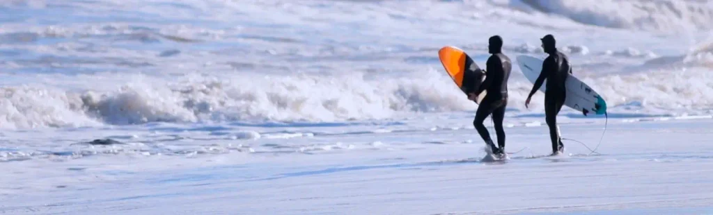 Two people in full black wetsuits walk away from the camera across wet sand, each carrying a surfboard (one orange-tipped, one white). Large foamy waves crash in the background.