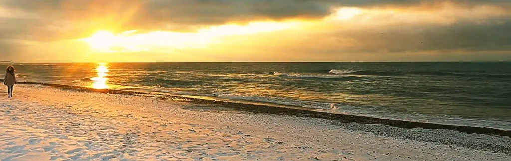 A person in a light coat stands on a snowy beach facing a bright golden sunrise or sunset over the ocean. The sun reflects on the water, with clouds illuminated in yellow and orange hues.