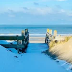 Wooden boardwalk and stairs lead through snow-dusted sand dunes toward the ocean. The path is framed by grassy dunes on both sides under a bright blue sky with scattered clouds.