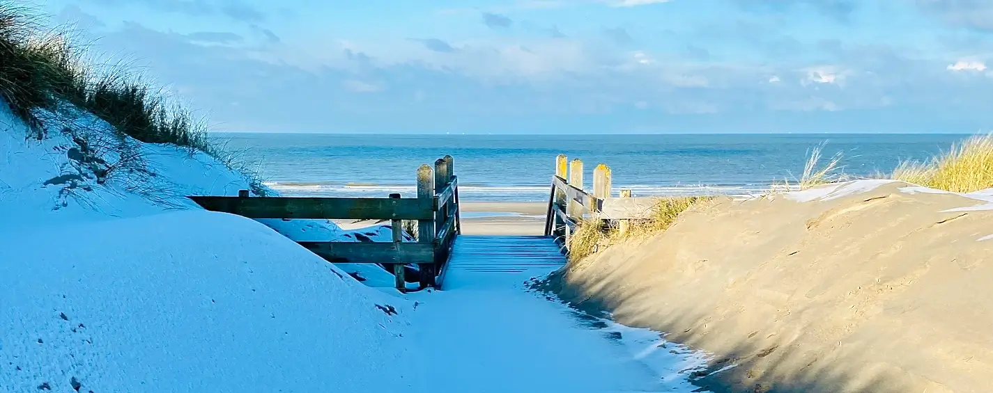 Wooden boardwalk and stairs lead through snow-dusted sand dunes toward the ocean. The path is framed by grassy dunes on both sides under a bright blue sky with scattered clouds.