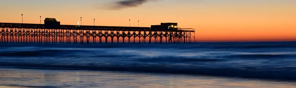 Silhouette of a long wooden pier extending into the ocean at sunset, with buildings and lights along the pier. The sky gradients from orange to yellow, while the foreground shows blue waves washing onto the wet sand.