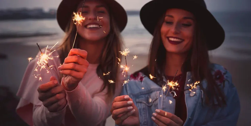 Two smiling young women on a beach at dusk hold lit sparklers. The woman on the left wears a brown hat and light top; the woman on the right wears a black hat, denim jacket, and has dark lipstick. Sparks fly from the sparklers against a soft evening sky and water background.

