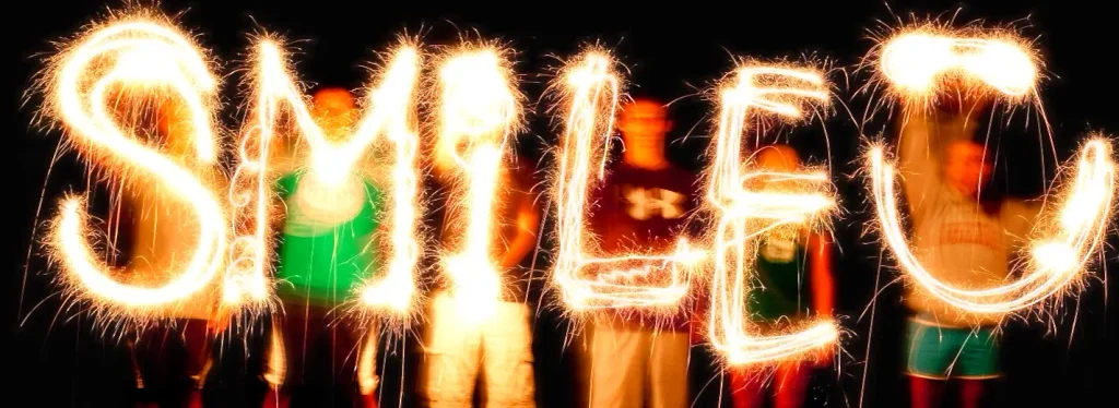 A long-exposure photo of several people standing in the dark, using sparklers to write the glowing orange word “SMILE” followed by a smiley face “:)” in the air.

