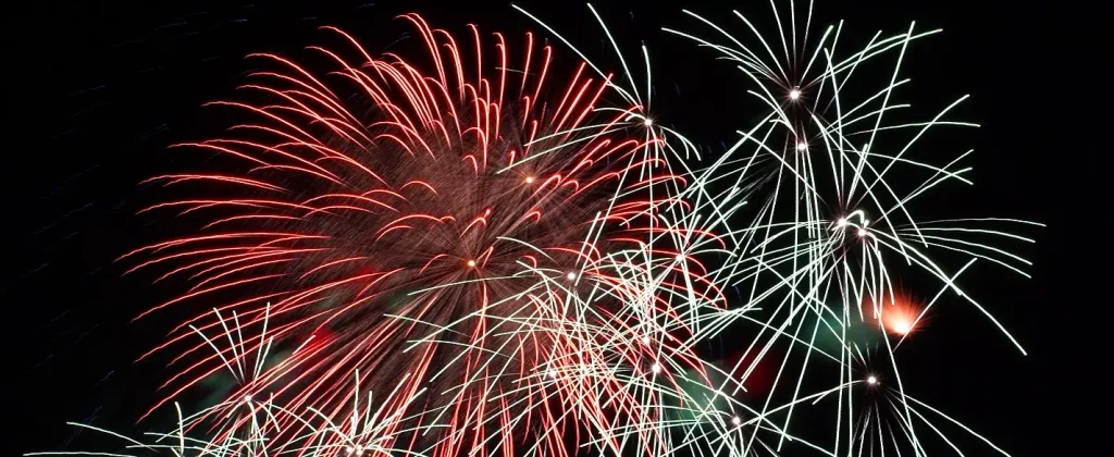 Nighttime fireworks with a large red starburst on the left and white/silver trailing bursts and streaks on the right against a black sky.

