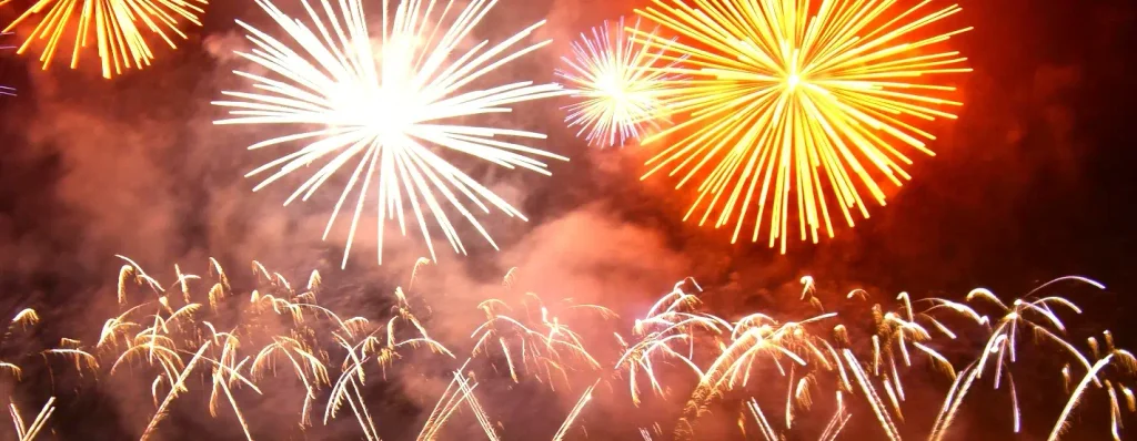 Bright yellow and white firework explosions high in a reddish night sky, with many golden trailing sparks and smoke rising from below.

