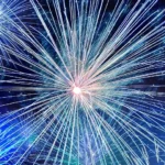 A vibrant nighttime fireworks display with large bursts in pink, purple, green, white, and blue exploding against a black sky. A bright white-yellow core is visible near the bottom center.