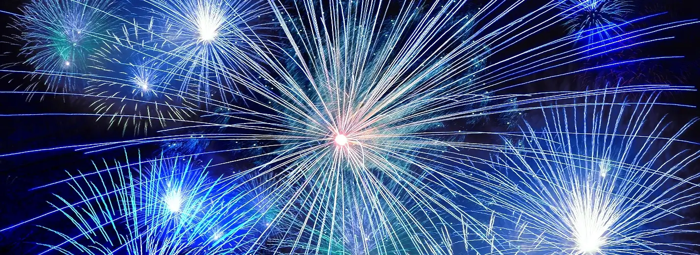 A vibrant nighttime fireworks display with large bursts in pink, purple, green, white, and blue exploding against a black sky. A bright white-yellow core is visible near the bottom center.